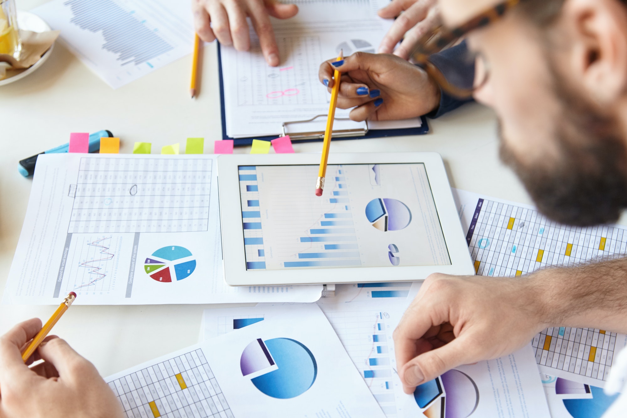 Bearded man in glasses analysing charts and graphics on touchpad with his colleagues. Young people w